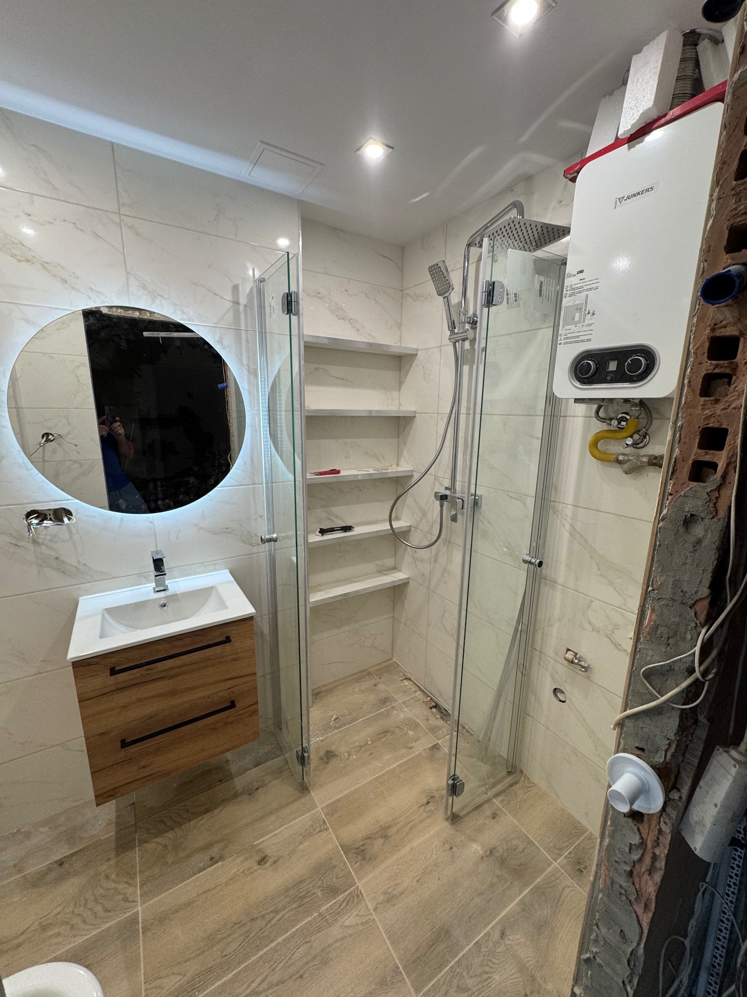 Wykończona łazienka z kabiną prysznicową, umywalką z szafką i okrągłym lustrem z podświetleniem LED. Widoczny bojler i półki w ścianie. Płytki imitujące marmur.