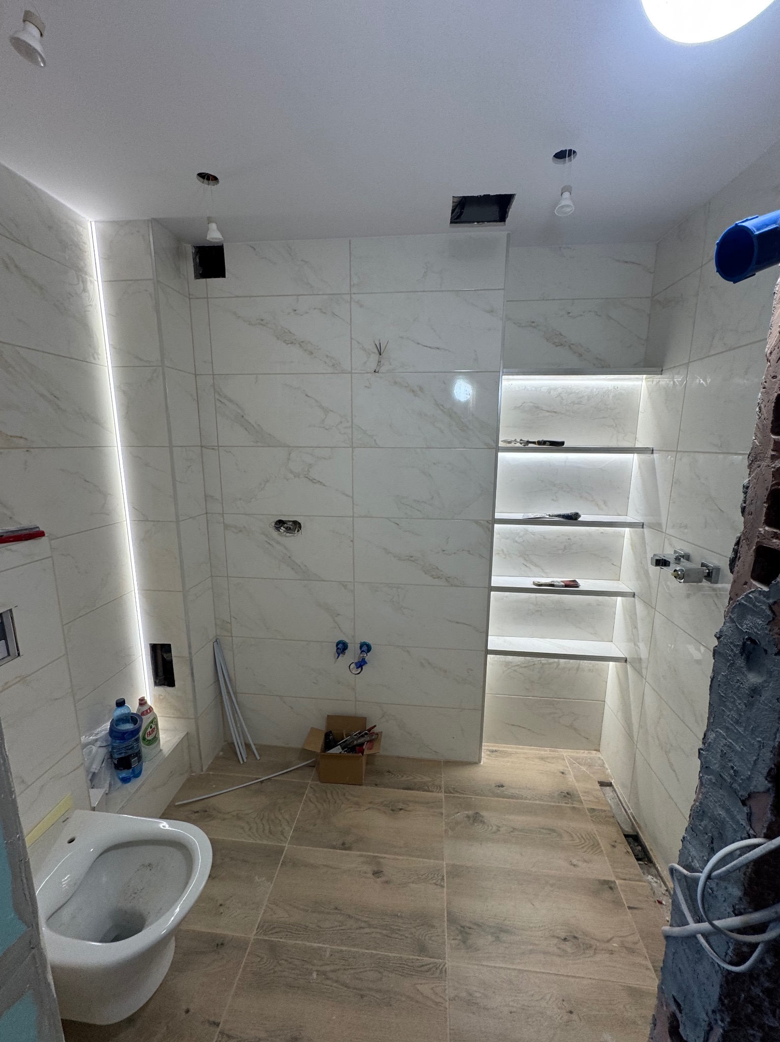 Łazienka w trakcie wykończenia: białe płytki imitujące marmur, drewnopodobna podłoga, wnęka z półkami podświetlona LED, widoczne narzędzia i materiały budowlane.