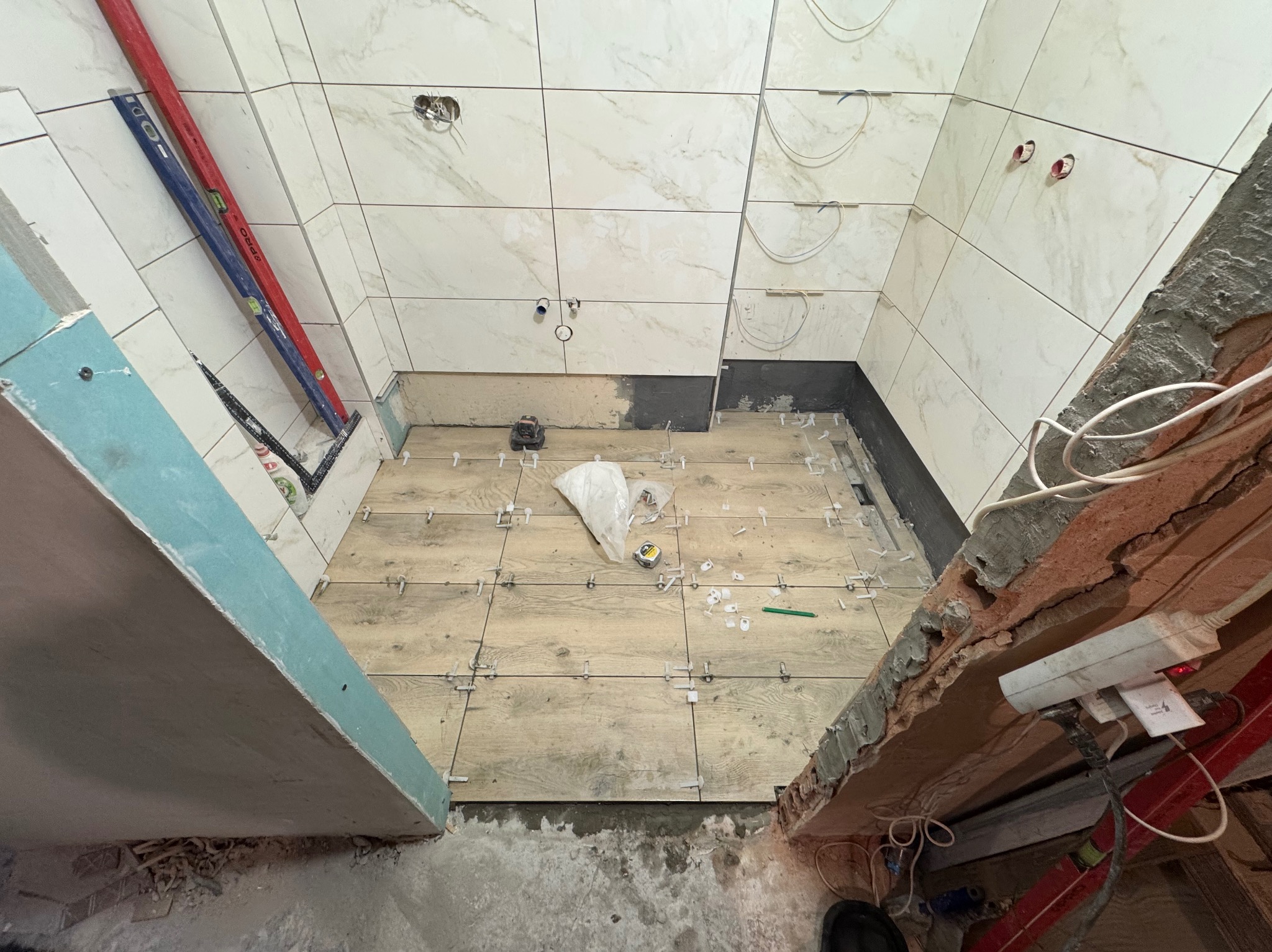 Układanie płytek podłogowych imitujących drewno w łazience, z krzyżykami dystansowymi. Widoczne płytki ścienne, poziomice i narzędzia glazurnicze. Prace wykończeniowe w toku.