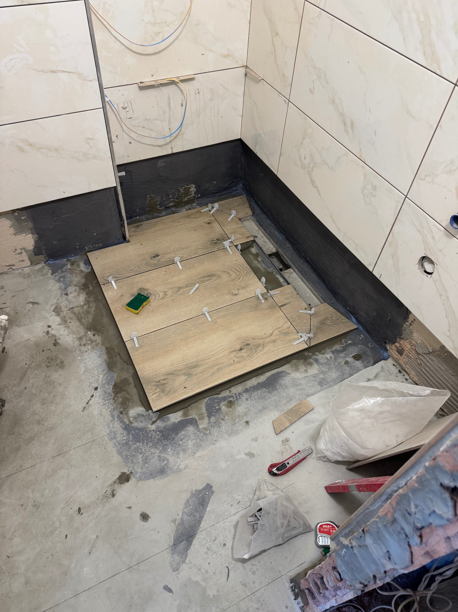 Układanie drewnopodobnych płytek podłogowych w łazience, widoczne krzyżyki dystansowe i gąbka do czyszczenia. Ściany wyłożone marmurkową glazurą.