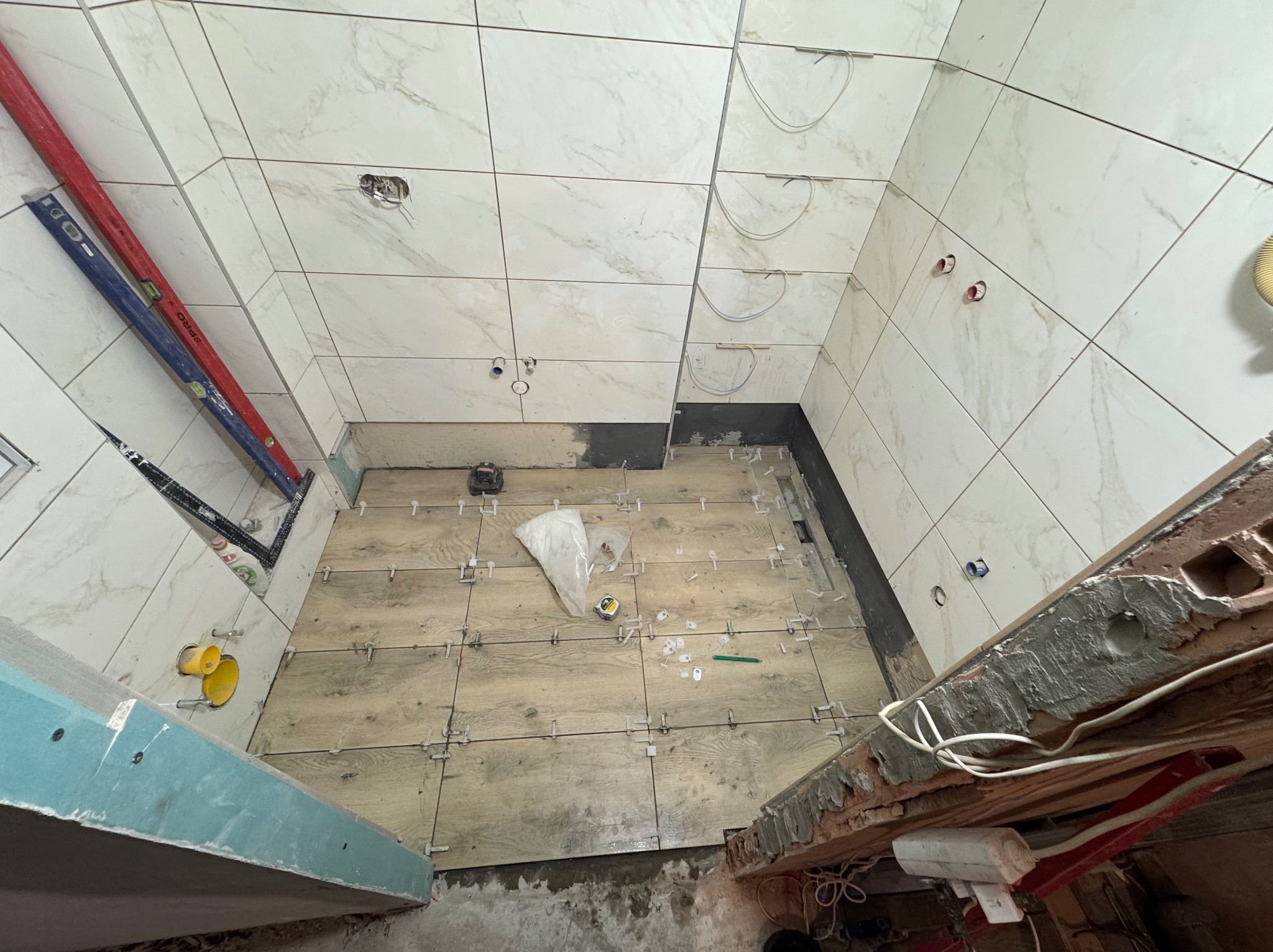 Wnętrze łazienki w trakcie prac glazurniczych: białe płytki marmurowe na ścianach, drewnopodobne płytki na podłodze, poziomice, krzyżyki dystansowe, widoczne instalacje.