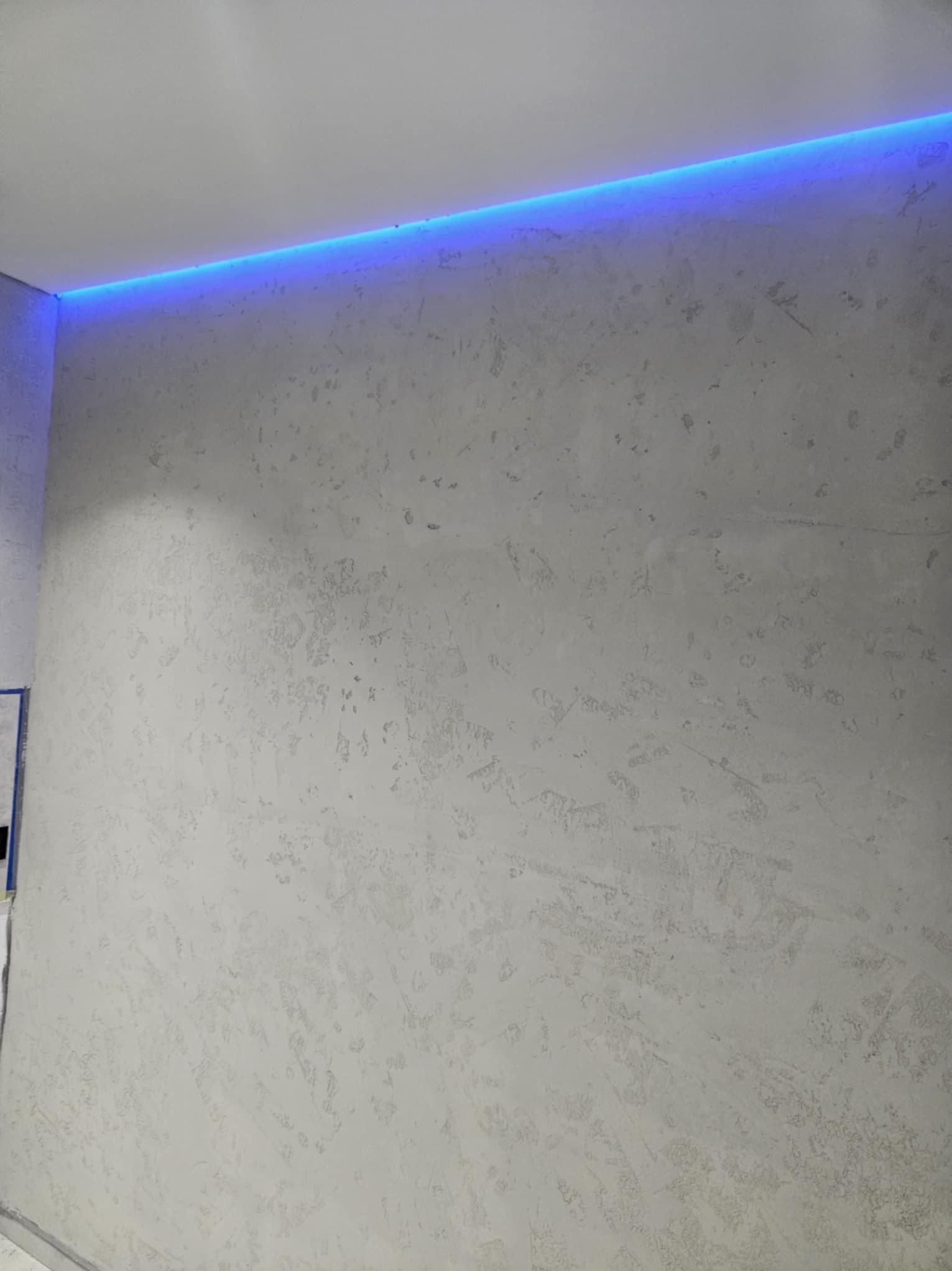 Ściana z dekoracyjną fakturą i subtelnym, niebieskim oświetleniem LED przy suficie. Minimalistyczny design wnętrza, neutralna kolorystyka.