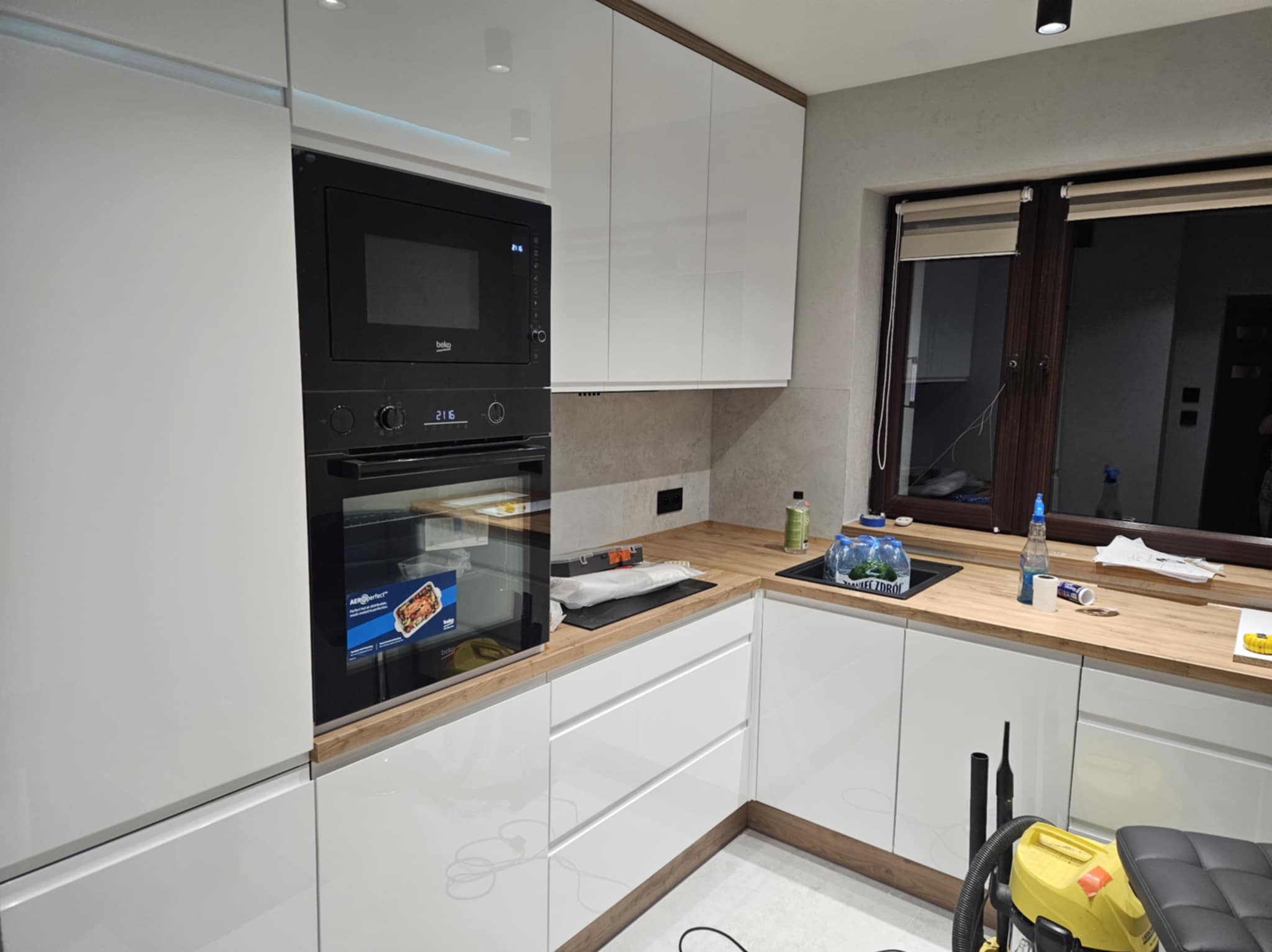 Nowoczesna, biała kuchnia z drewnianym blatem i wbudowanym piekarnikiem Beko. Widoczne okno z roletami i sprzęt czyszczący. Minimalistyczny design.