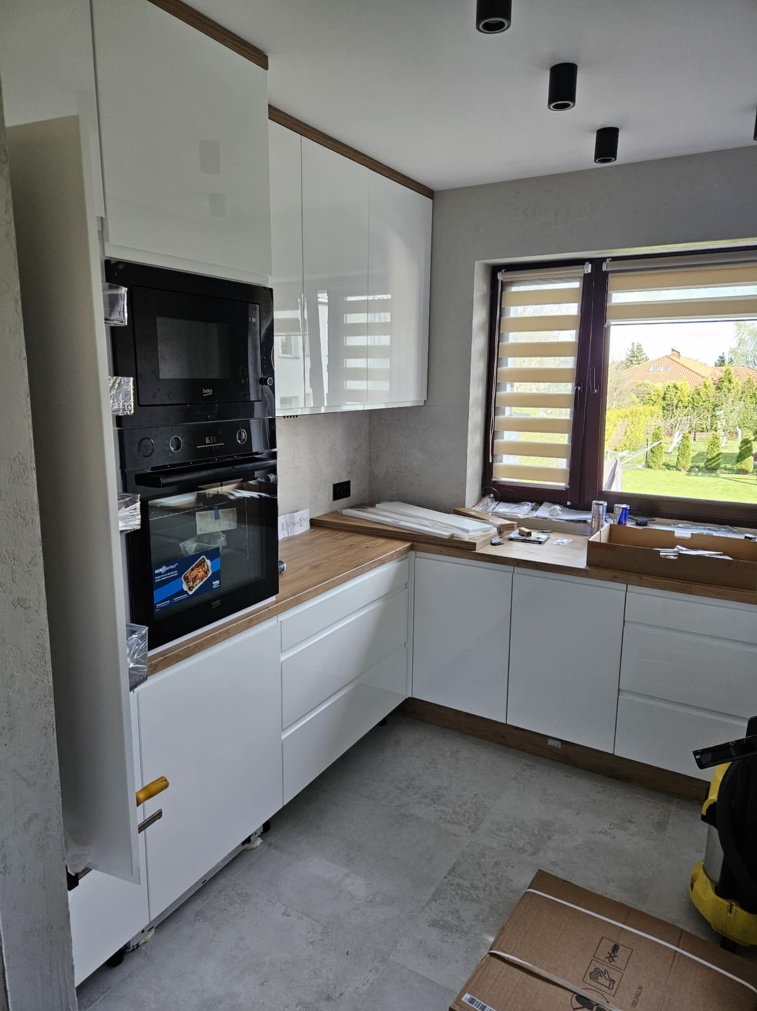 Montaż białej, nowoczesnej kuchni z drewnianym blatem. Widoczne zabudowane sprzęty AGD i okno z roletami. Częściowo widoczne kartony i narzędzia.