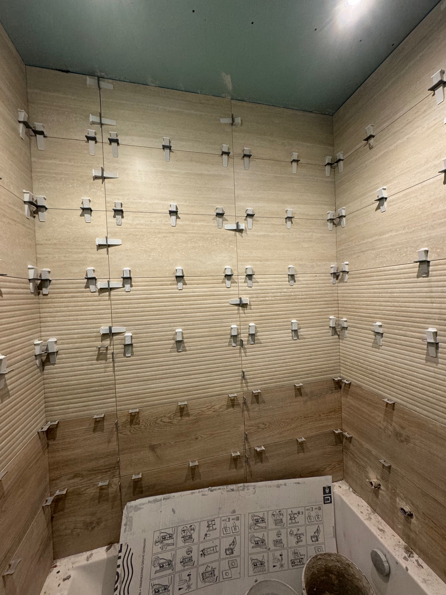 Układanie beżowych płytek imitujących drewno w łazience, z użyciem krzyżyków dystansowych i sznurka do poziomowania. Widoczna wanna i karton po płytkach.