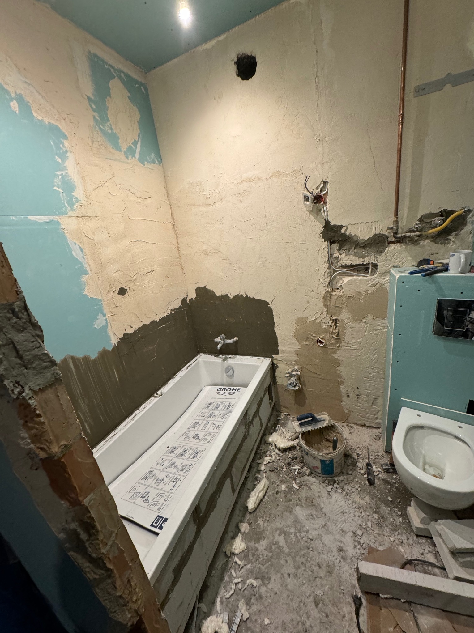 Łazienka w trakcie remontu: surowe ściany z nałożoną gładzią, nowa wanna Grohe obudowana cegłami, zamontowana toaleta, widoczne instalacje i narzędzia.