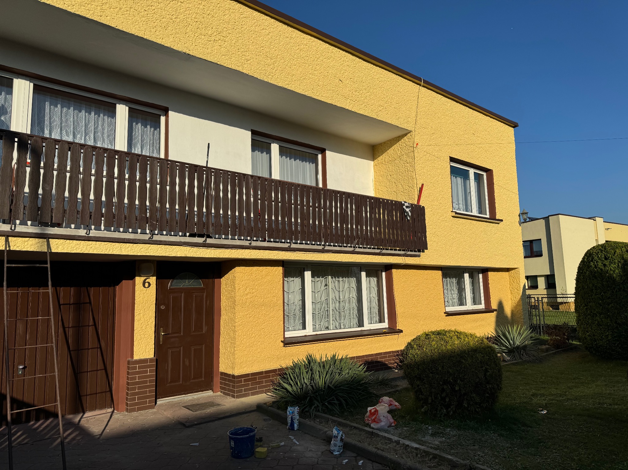 Żółta elewacja dwupiętrowego domu z balkonem, brązowe okna i drzwi. Widoczne ślady farby i narzędzia malarskie, sugerujące świeżo zakończone malowanie.