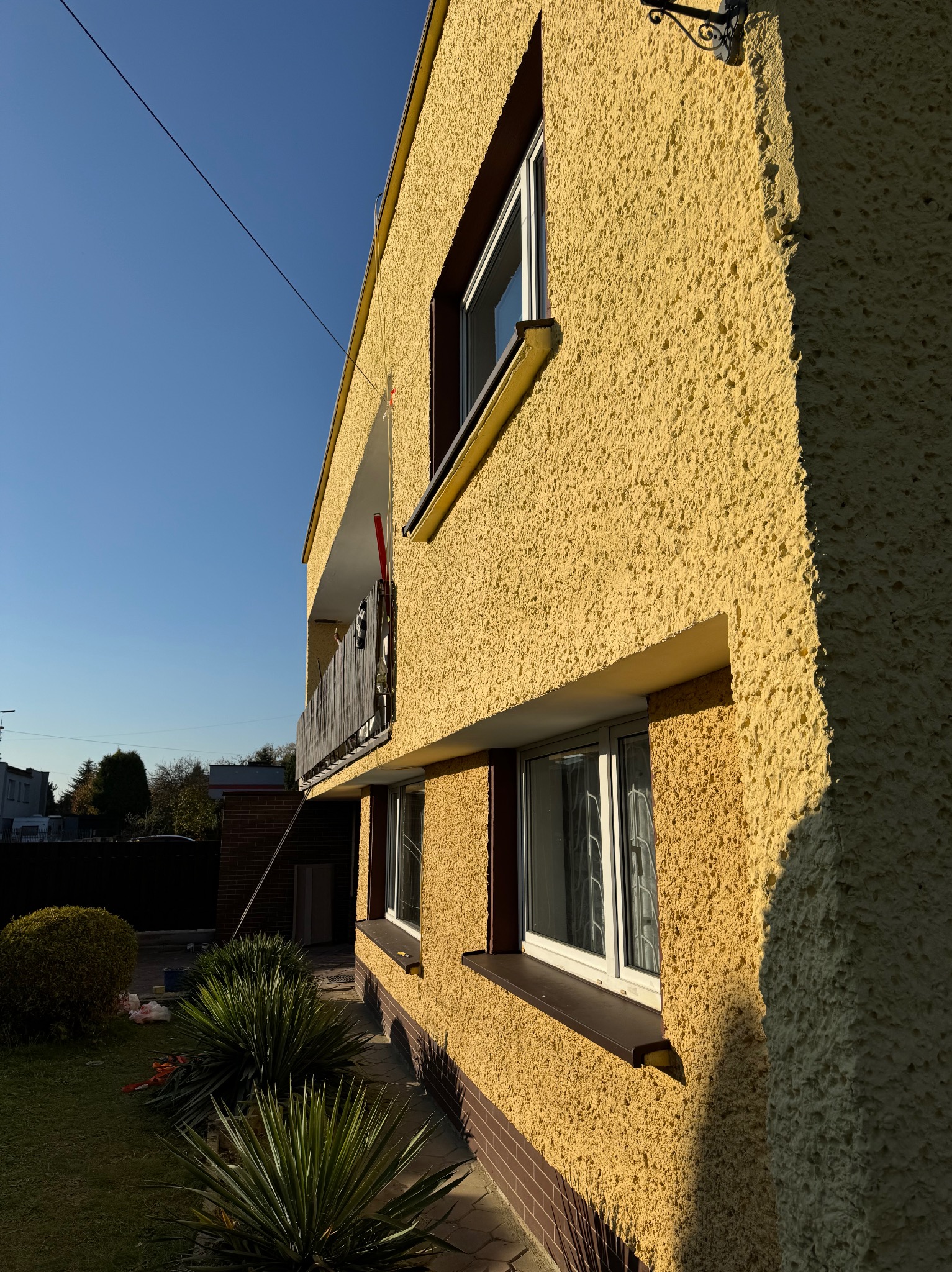 Żółta elewacja dwupiętrowego domu z balkonem, widoczna z boku w słoneczny dzień. Charakterystyczna faktura tynku i ciemne obramowania okien.