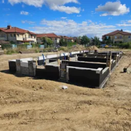 Fundamenty domu jednorodzinnego w trakcie budowy, z widoczną izolacją przeciwwilgociową i elementami szalunku, na tle błękitnego nieba z chmurami.