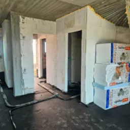 Wnętrze w trakcie budowy: ściany z białych bloczków, betonowy sufit, instalacja podłogowa i stos styropianu Terma Organika, widoczne ślady robocze.