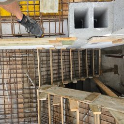 Biankowscy spolka z o.o. - Zbrojenie fundamentów z widoczną siatką stalową, szalunkiem drewnianym i fragmentem buta pracownika. Widok z góry na plac budowy.