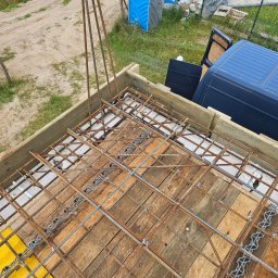 Biankowscy spolka z o.o. - Zbrojenie fundamentu w szalunku z desek, widok z góry, Miechucino. Elementy zbrojenia połączone drutem. Na zewnątrz budowy zaparkowany bus i toaleta.