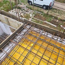 Biankowscy spolka z o.o. - Zbrojenie fundamentów z żółtą szalunkową płytą, widoczne drewniane deski szalunkowe i fragment białego busa w tle. Ujęcie z góry, z widoczną siatką zbrojeniową.