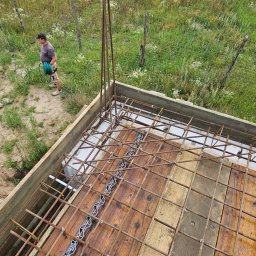 Biankowscy spolka z o.o. - Zbrojenie fundamentu domu z drewnianym szalunkiem, widok z góry. W tle mężczyzna z wężem ogrodowym i zielona trawa. Elementy zbrojenia połączone drutem.