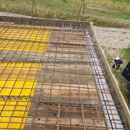 Biankowscy spolka z o.o. - Zbrojenie płyty fundamentowej z żółtymi szalunkami i wystającymi prętami, widok z góry na budowę. W tle zieleń i pracownik przy krawędzi.