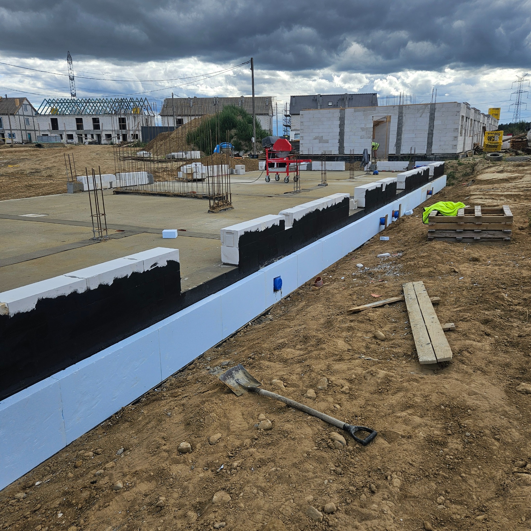 Fundamenty domu w trakcie izolacji: biały styropian, czarna izolacja. W tle budowa, szpadel na pierwszym planie. Pochmurne niebo.