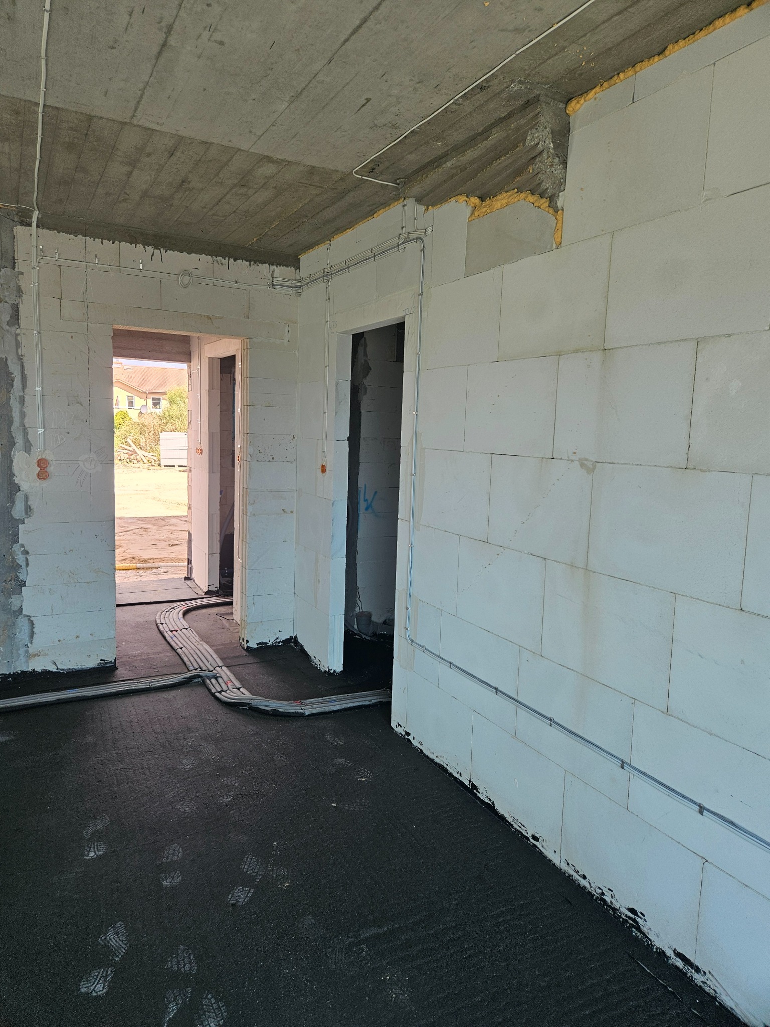 Wnętrze w trakcie budowy: ściany z betonu komórkowego, instalacje elektryczne w peszlach na podłodze pokrytej czarną membraną, widoczne ślady butów, surowy betonowy strop.