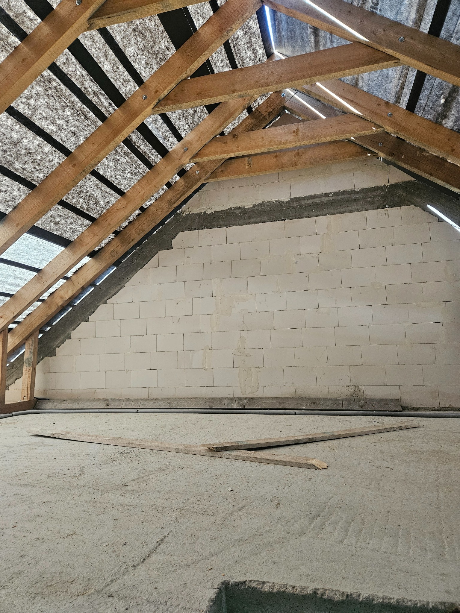 Surowy stan poddasza: bloczki, drewniana więźba dachowa z membraną, betonowa wylewka. Fragment instalacji przy ścianie. Deski leżą na podłodze.