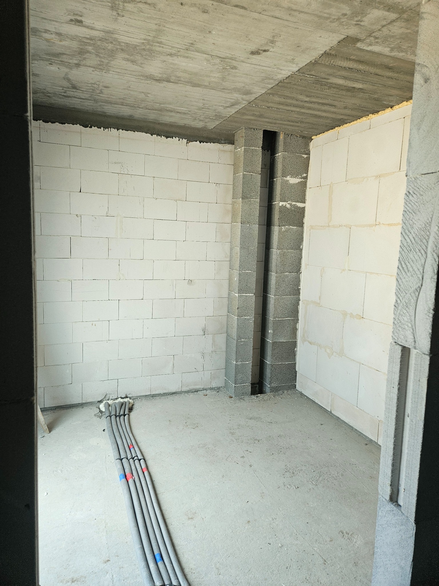 Wnętrze w stanie surowym: białe ściany z betonu komórkowego, betonowy sufit, piony instalacyjne z szarego betonu i wiązka szarych rur instalacyjnych na podłodze.