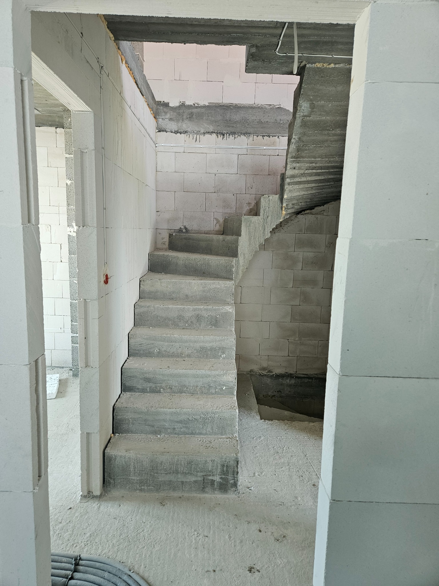 Surowe schody betonowe w trakcie budowy wewnątrz budynku z białych bloczków. Widoczny fragment instalacji elektrycznej. Kadr z dołu, surowy stan.