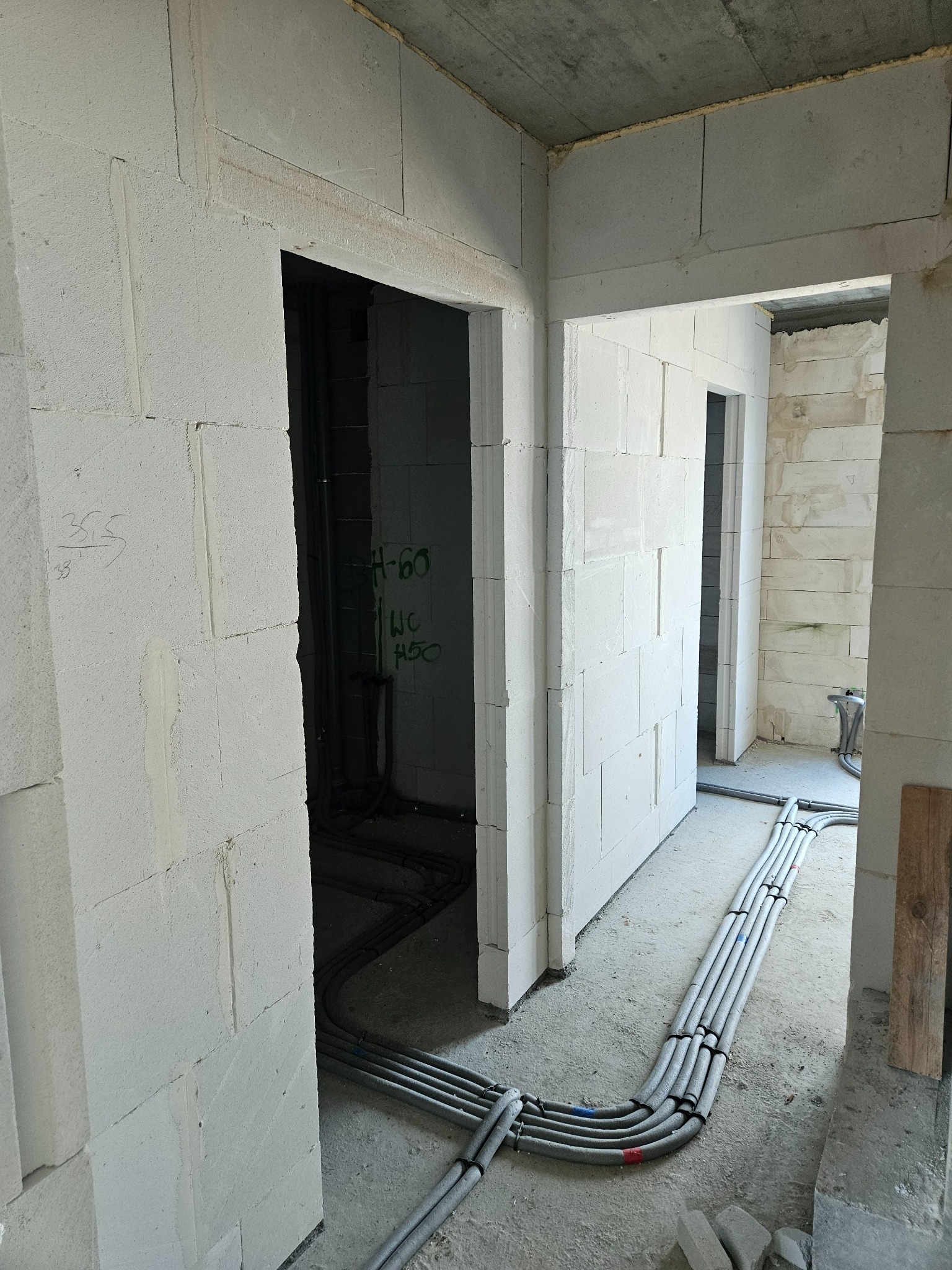 Wnętrze w trakcie budowy: ściany z betonu komórkowego, instalacje elektryczne ułożone na podłodze, widoczne otwory drzwiowe i napisy na ścianach.
