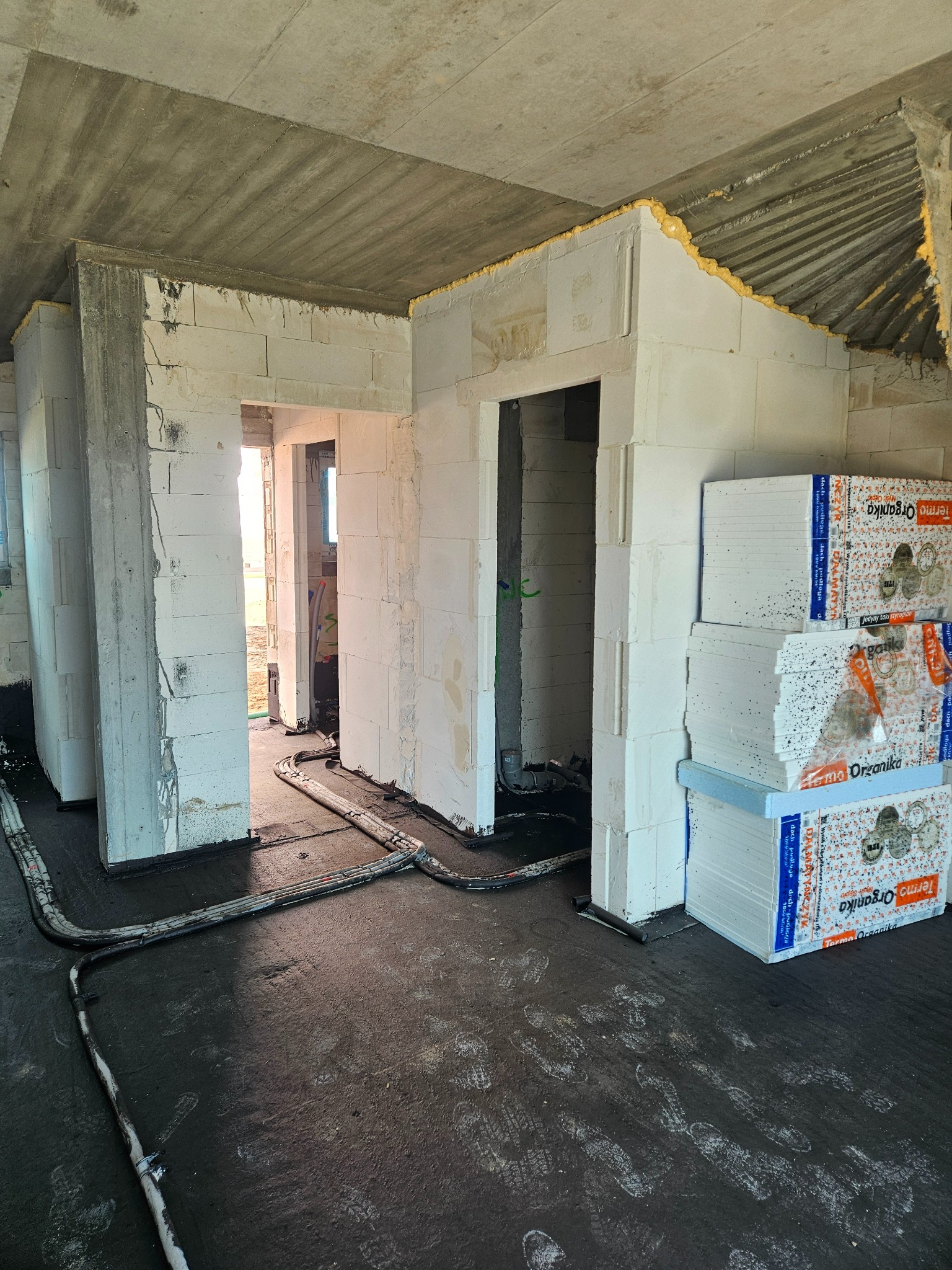 Wnętrze w trakcie budowy: ściany z białych bloczków, betonowy sufit, instalacja podłogowa i stos styropianu Terma Organika, widoczne ślady robocze.