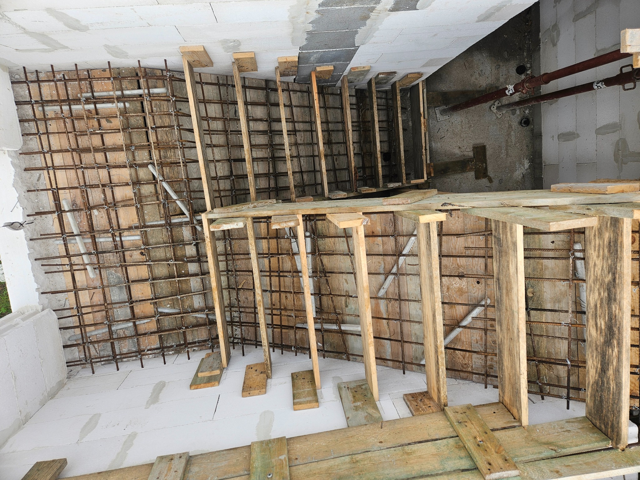 Zbrojenie ściany z drewnianym szalunkiem i instalacją sanitarną w surowym wnętrzu budynku. Widok z góry na etap przygotowawczy przed betonowaniem.