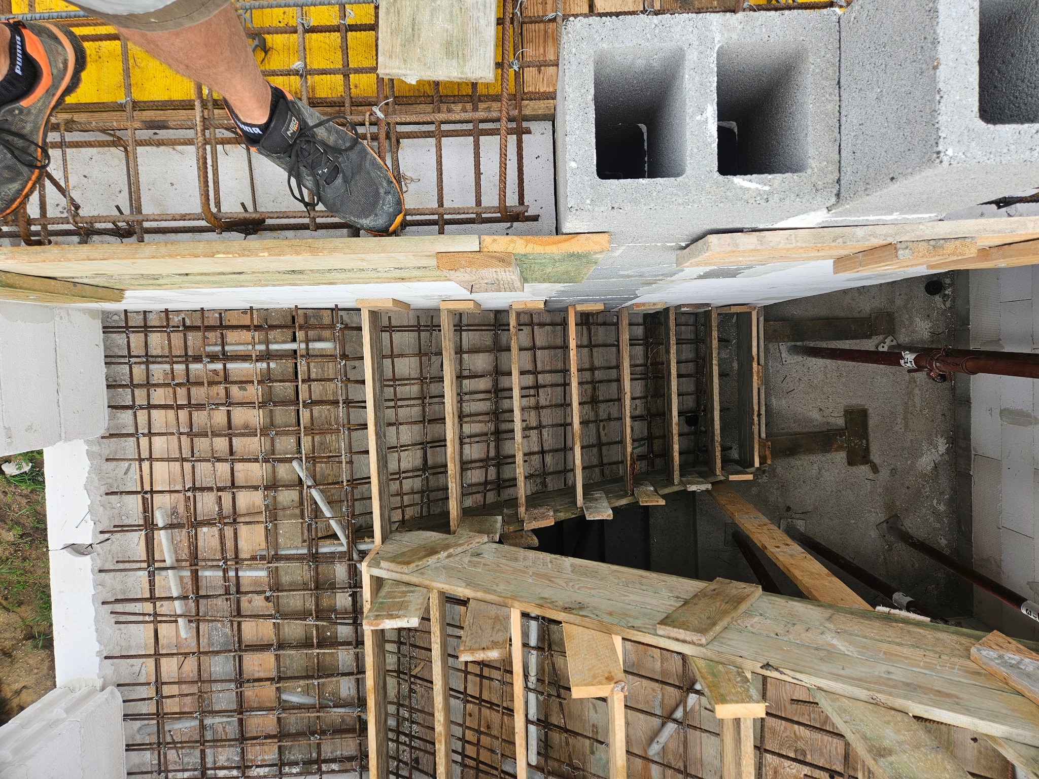 Zbrojenie fundamentów z widoczną siatką stalową, szalunkiem drewnianym i fragmentem buta pracownika. Widok z góry na plac budowy.