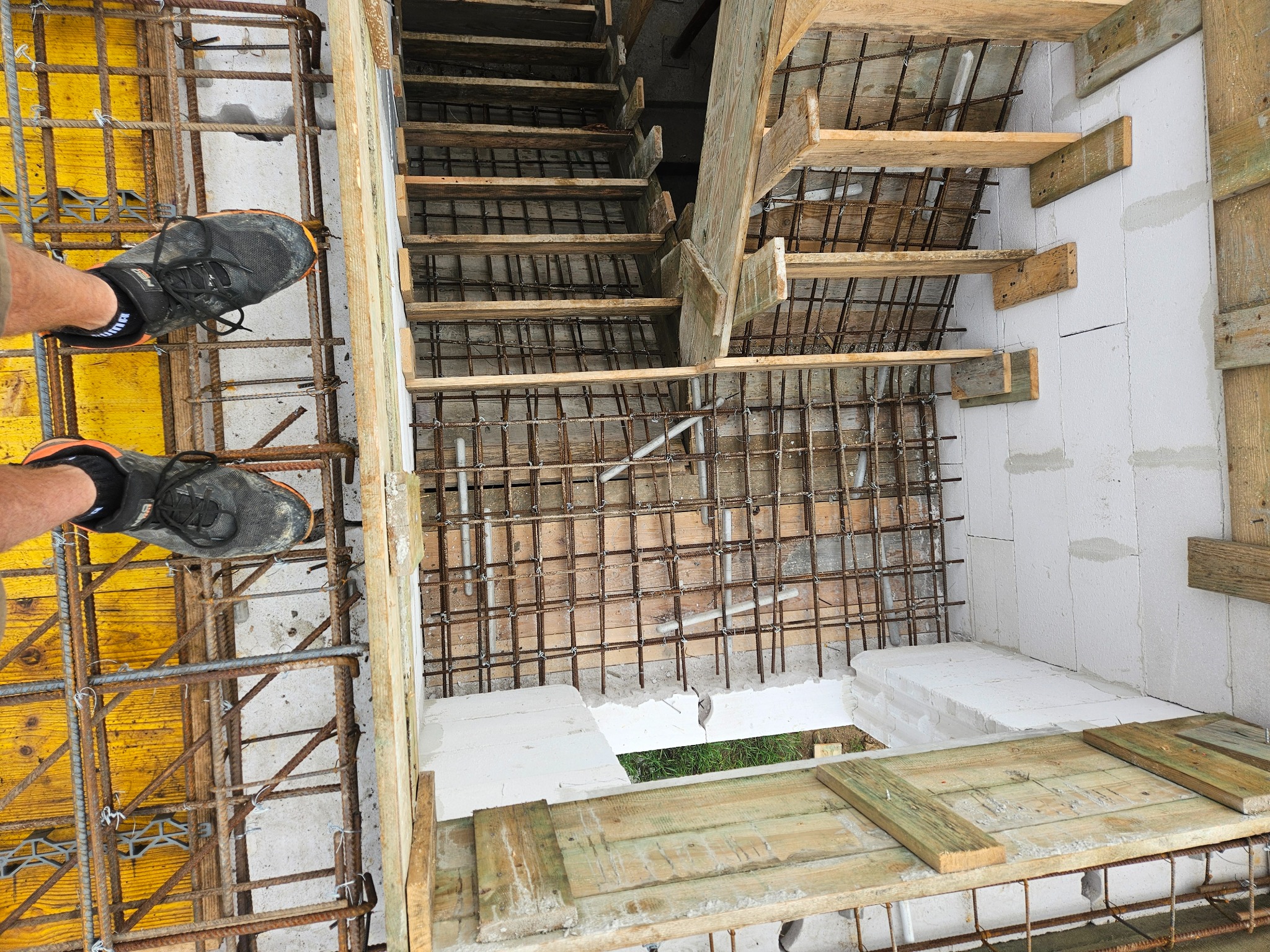 Zbrojenie schodów i fundamentów z widocznymi stopami pracownika w obuwiu roboczym na żółtym szalunku. Ujęcie z góry ukazuje etap budowy.