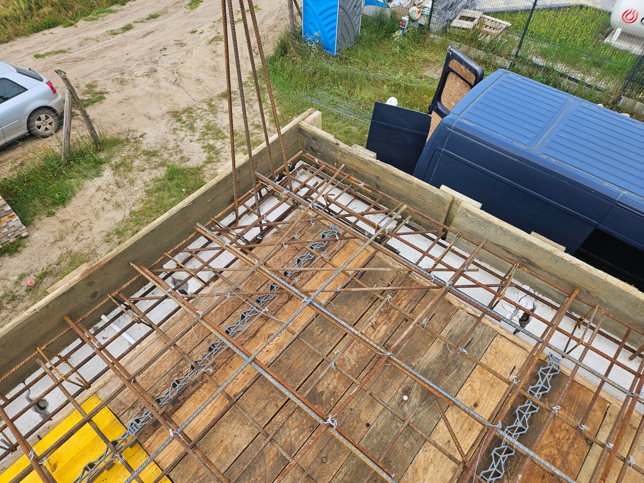 Zbrojenie fundamentu w szalunku z desek, widok z góry, Miechucino. Elementy zbrojenia połączone drutem. Na zewnątrz budowy zaparkowany bus i toaleta.