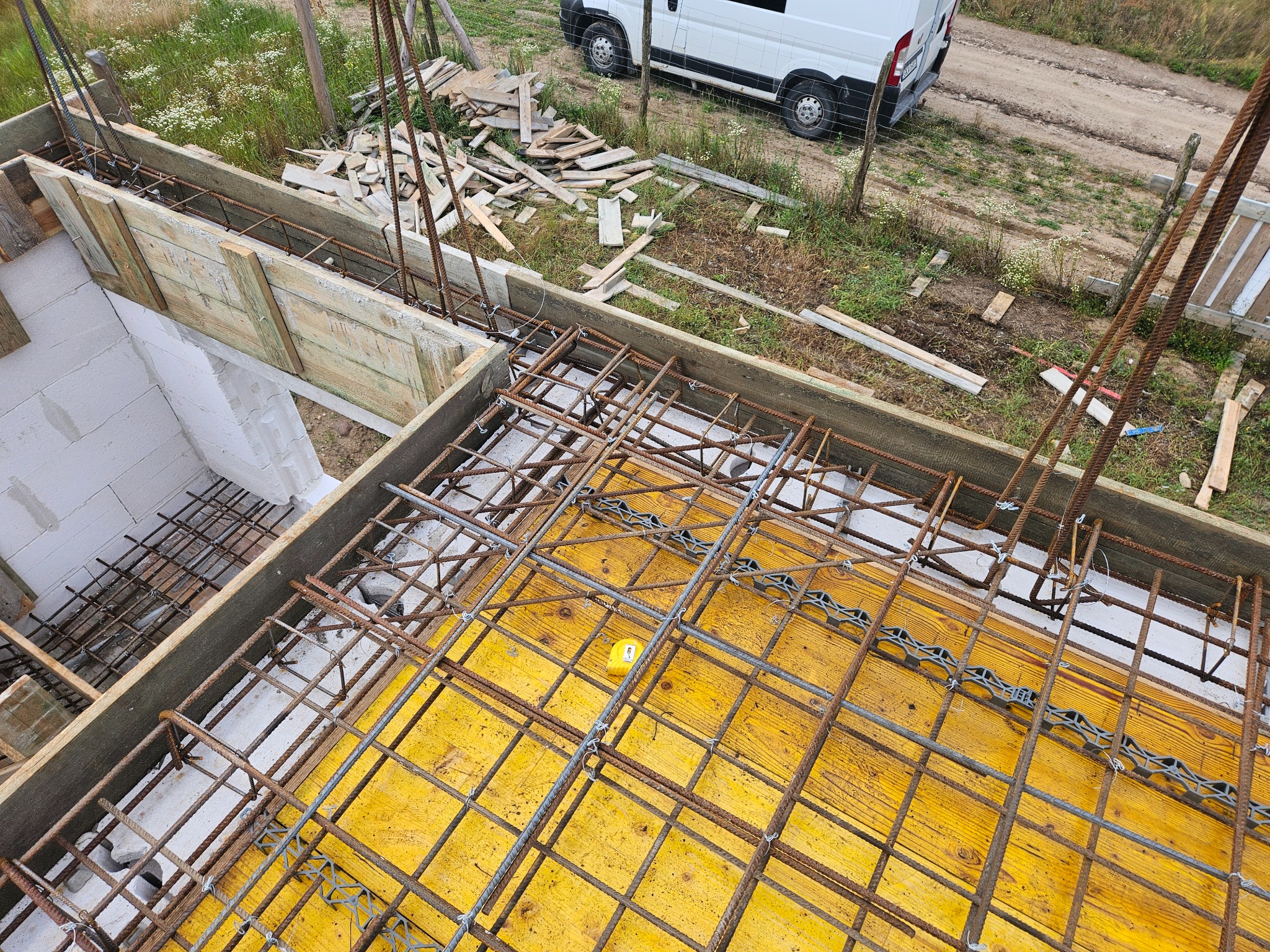 Zbrojenie fundamentów z żółtą szalunkową płytą, widoczne drewniane deski szalunkowe i fragment białego busa w tle. Ujęcie z góry, z widoczną siatką zbrojeniową.