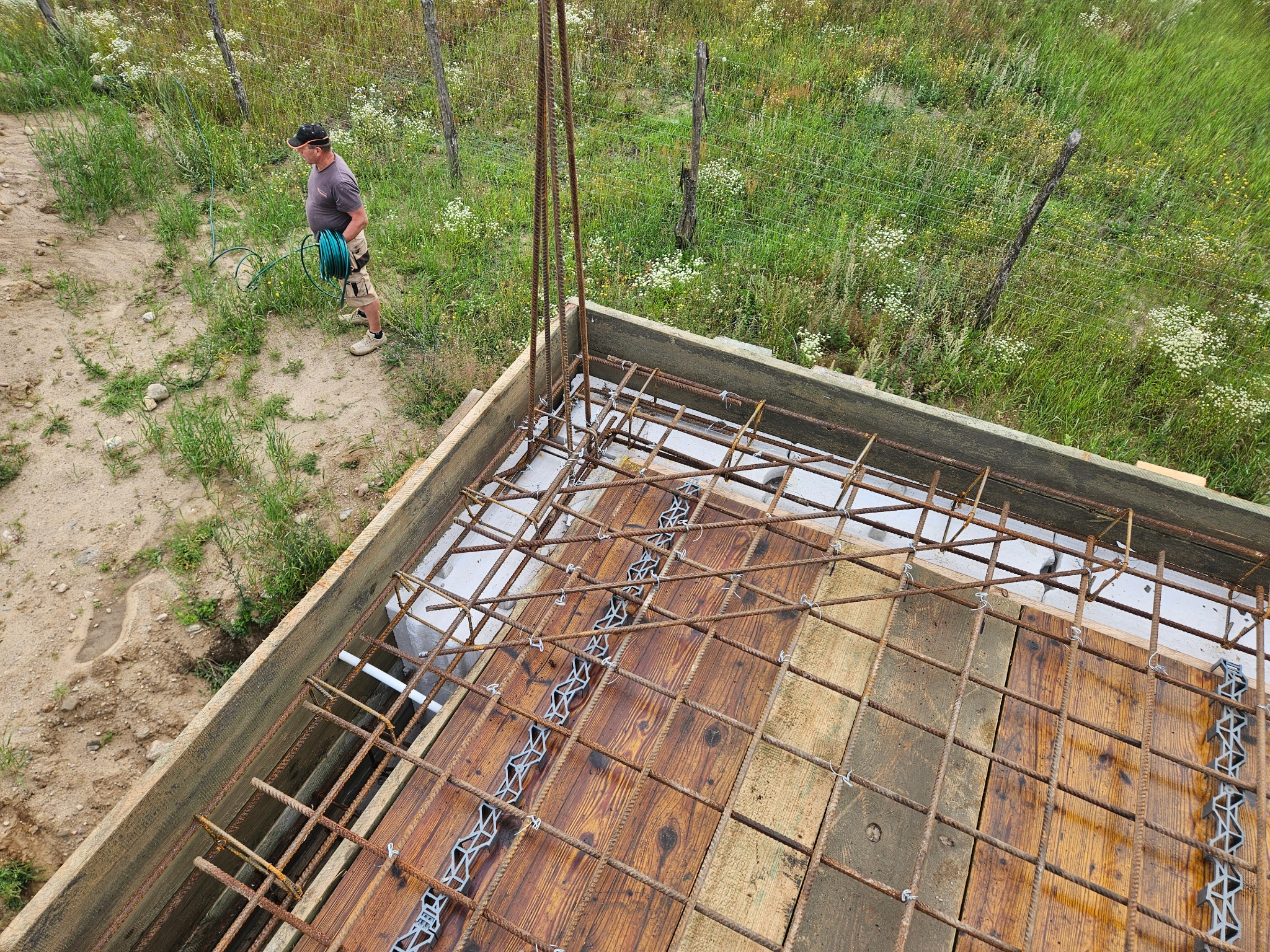 Zbrojenie fundamentu domu z drewnianym szalunkiem, widok z góry. W tle mężczyzna z wężem ogrodowym i zielona trawa. Elementy zbrojenia połączone drutem.