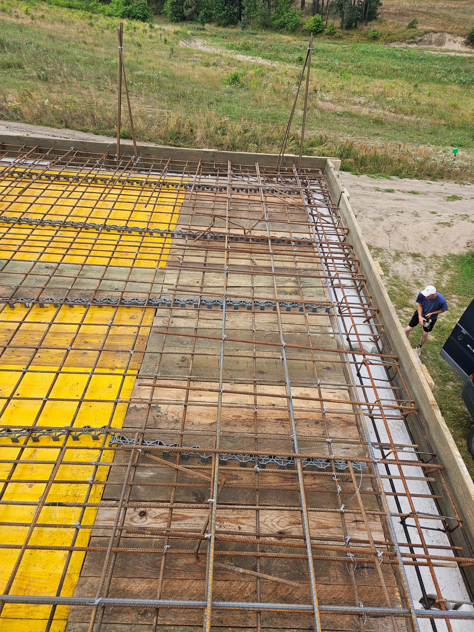 Zbrojenie płyty fundamentowej z żółtymi szalunkami i wystającymi prętami, widok z góry na budowę. W tle zieleń i pracownik przy krawędzi.
