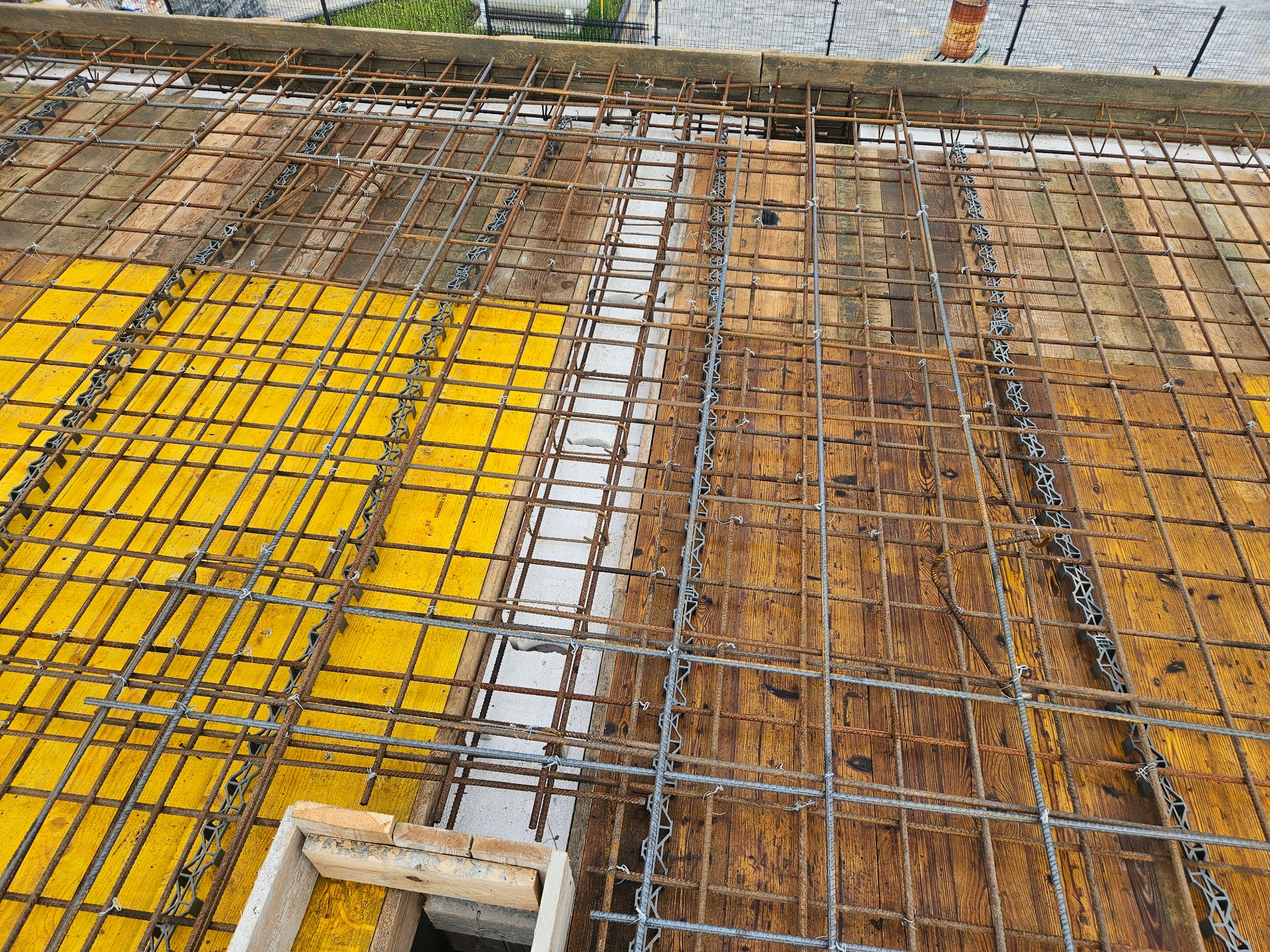 Zbrojenie stropu z siatki stalowej ułożonej na żółtym szalunku i deskach. Widoczne elementy dystansowe. Perspektywa z góry ukazuje etap budowy.