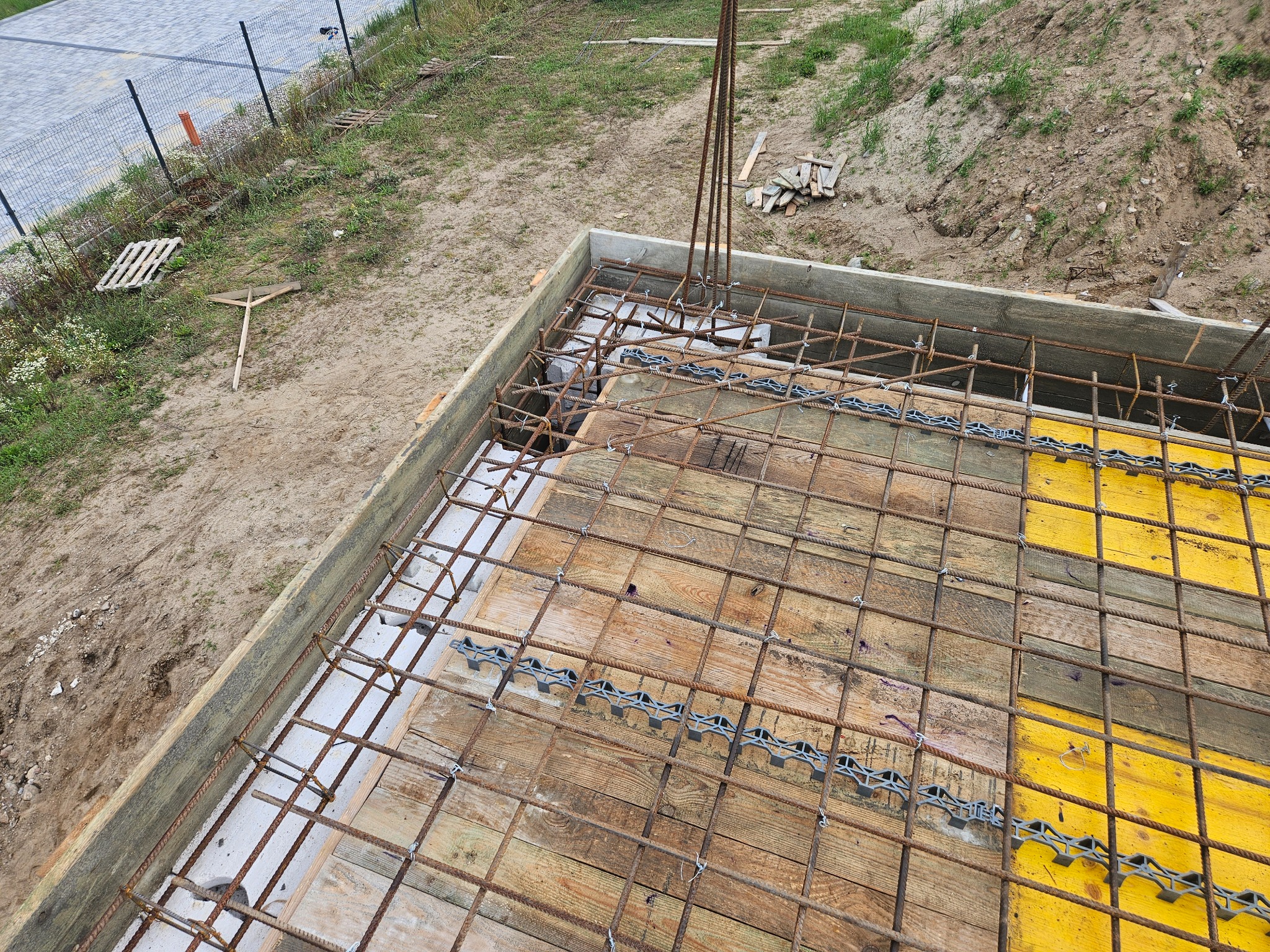 Zbrojenie fundamentu z drewnianym szalunkiem i elementami dystansowymi, widok z góry. Częściowo zalany beton. Na ziemi leżą narzędzia i paleta.