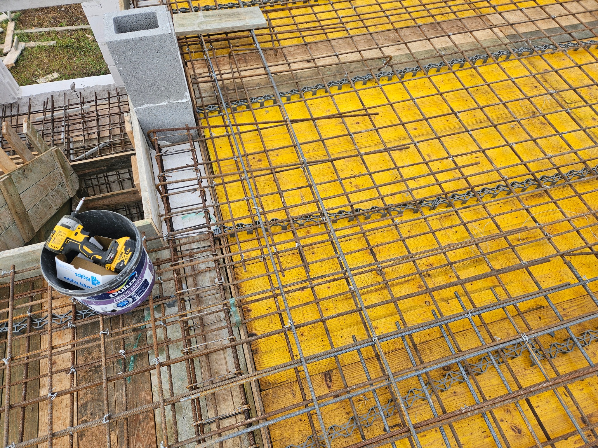 Zbrojenie stropu z żółtym szalunkiem, widoczne narzędzia w wiadrze. Detal budowy budynku żelbetowego z użyciem prętów i dystansów z tworzywa.