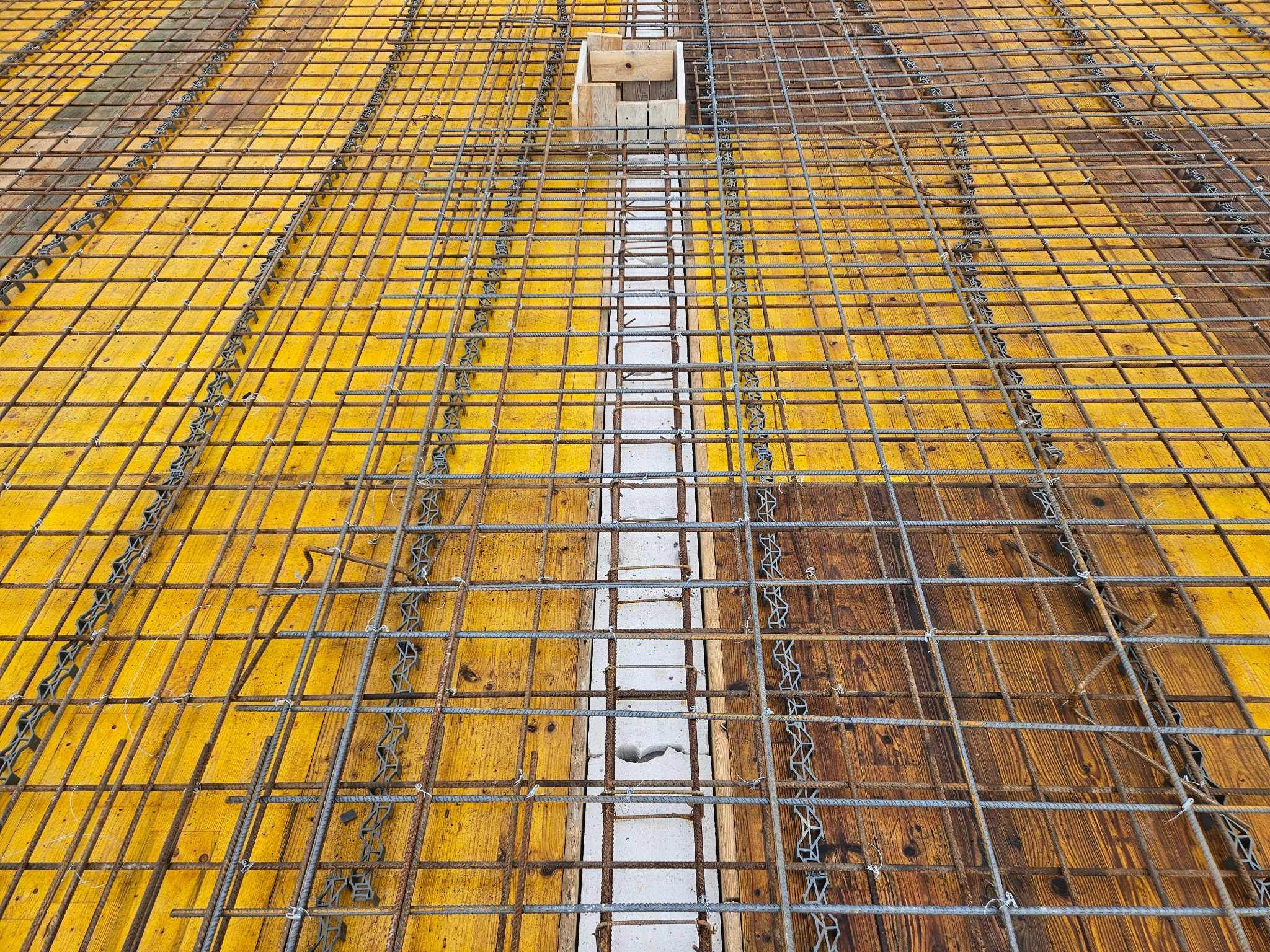 Zbrojenie płyty fundamentowej z żółtym szalunkiem i drewnianą formą, widok z góry. Metalowa siatka zbrojeniowa przygotowana do zalania betonem.