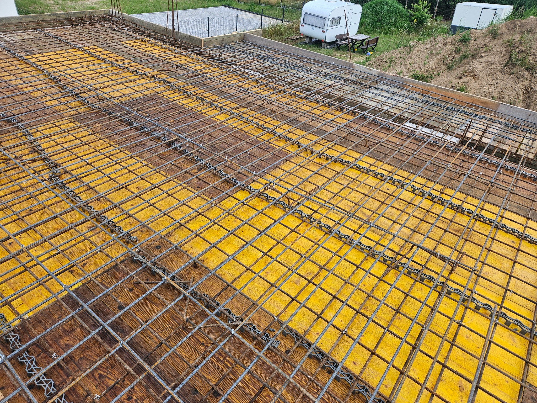 Zbrojenie stropu z widocznymi żółtymi deskami szalunkowymi i elementami dystansowymi. W tle zarys przyczepy kempingowej i zieleni. Prace budowlane w toku.