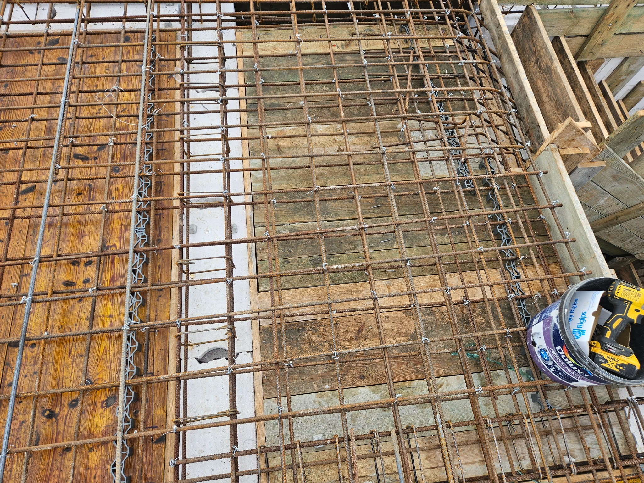 Zbrojenie szkieletu płyty fundamentowej z widocznymi drewnianymi szalunkami i narzędziem DeWalt w wiadrze farby Dulux, gotowe do zalania betonem.