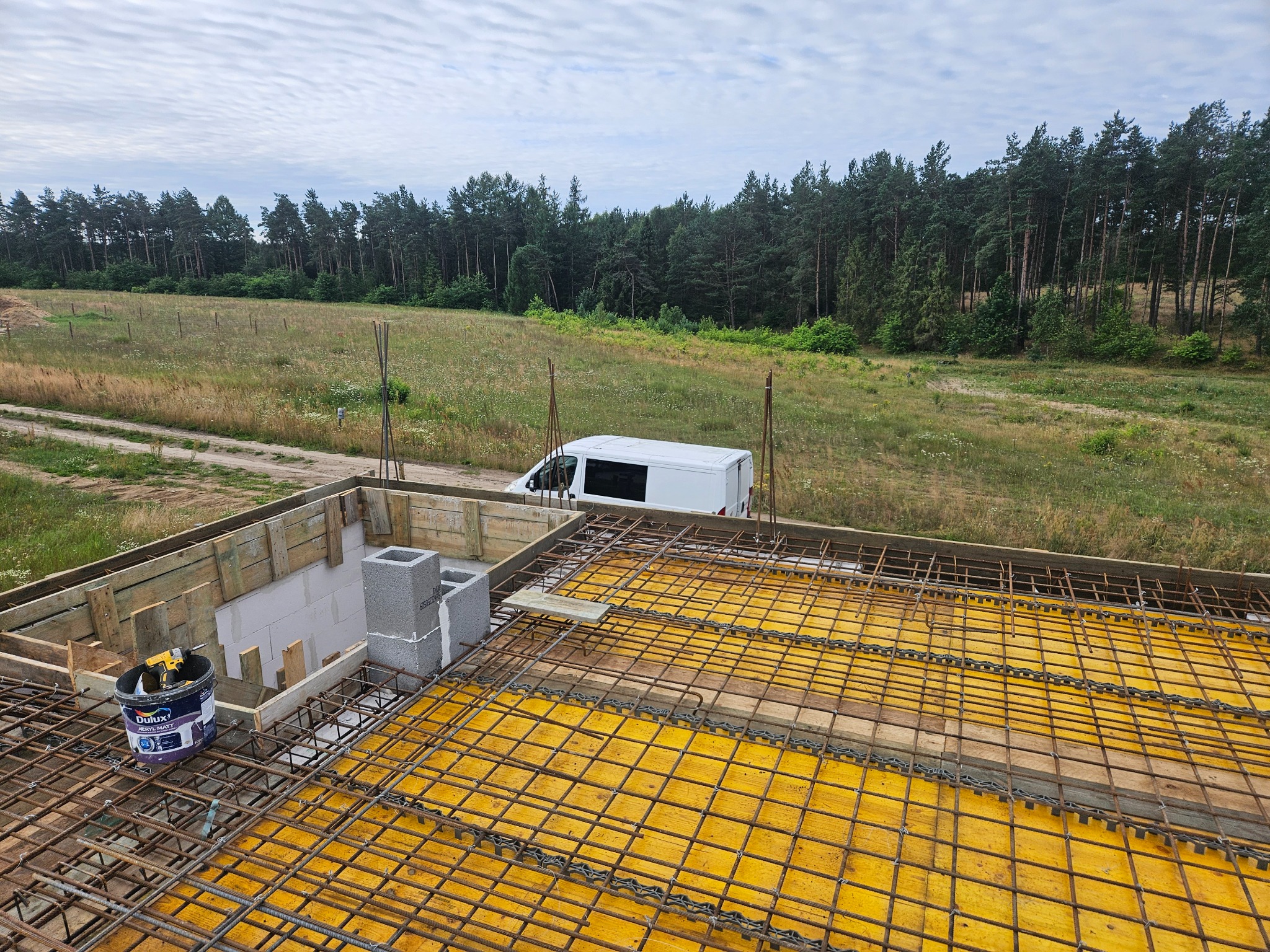 Zbrojenie stropu budynku żelbetowego na żółtym szalunku, widok z góry. W tle las i biały bus.