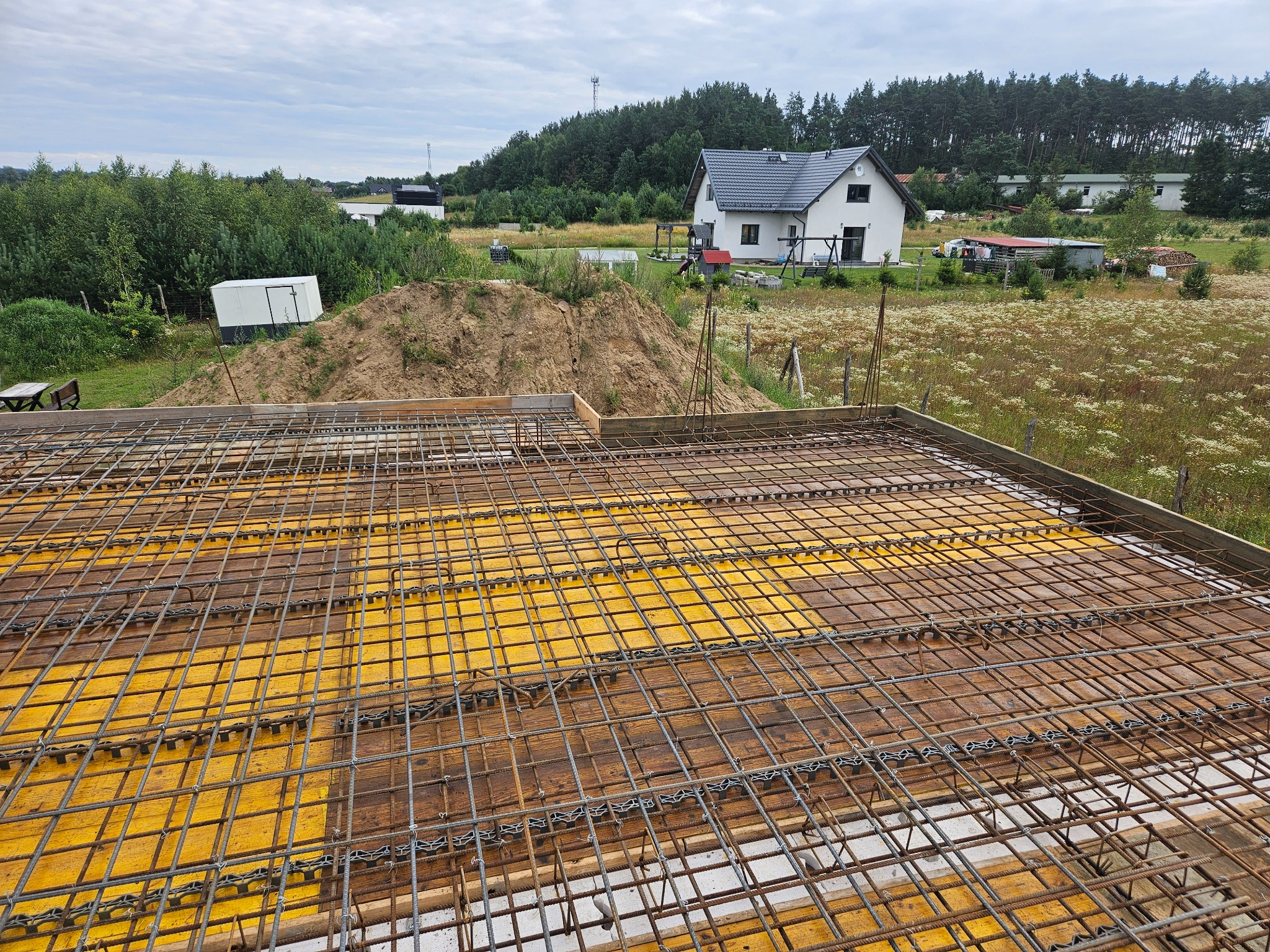 Zbrojenie stropu na żółtym szalunku, widok z góry. W tle dom jednorodzinny, drzewa i błękitne niebo. Prace budowlane w toku, etap przygotowania do wylania betonu.