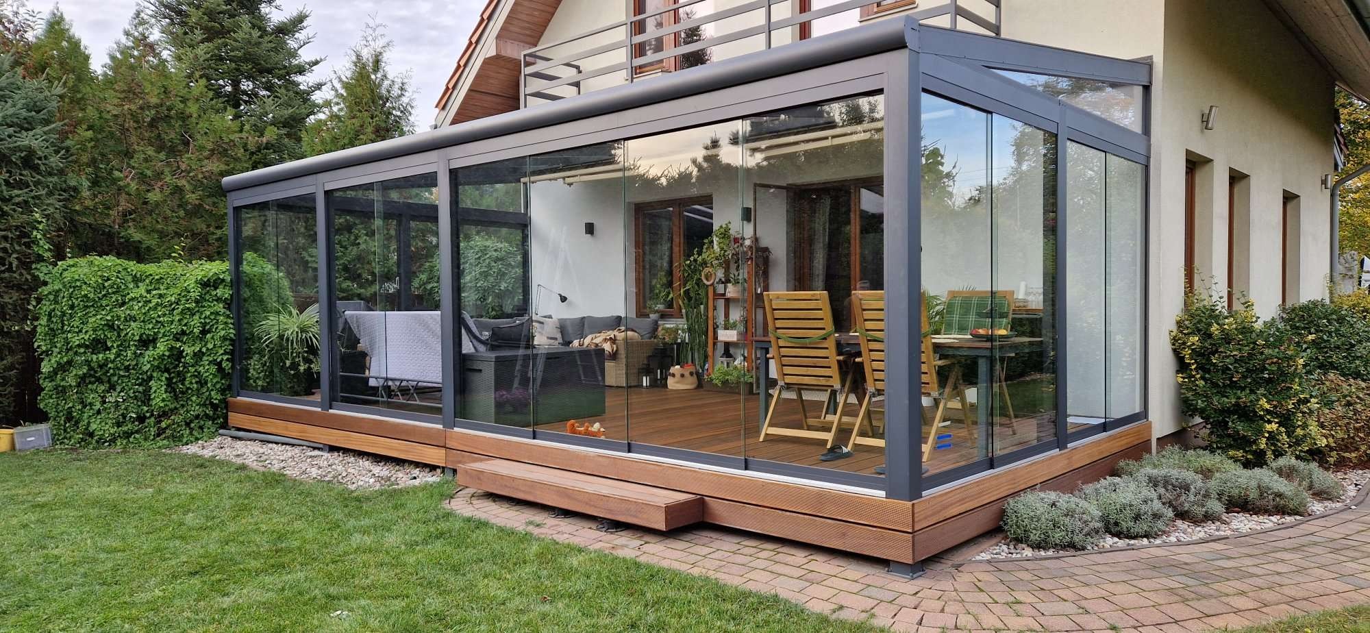 Zadaszony taras z drewnianą podłogą i szklanymi ścianami w ogrodzie. Wnętrze tarasu urządzone meblami wypoczynkowymi i stołem z krzesłami.