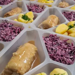 Porcje rolad z sosem, ziemniaków i surówki z czerwonej kapusty w plastikowych pojemnikach. Gotowe dania cateringowe, ułożone w rzędach na stole.