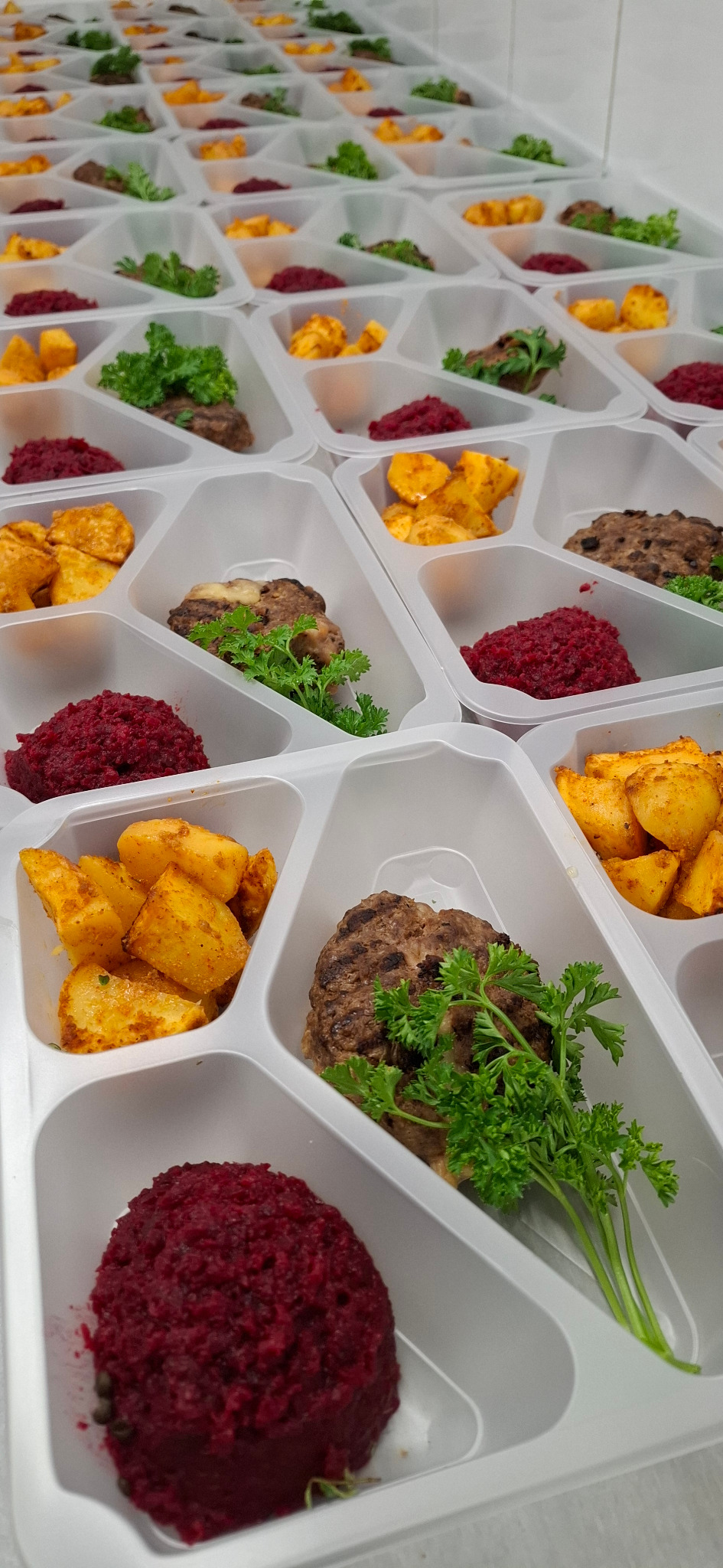 Dietetyczne posiłki w białych tackach z trzema przegródkami, zawierające kotlet, buraczki i ziemniaki, gotowe do wydania w ramach cateringu dietetycznego.