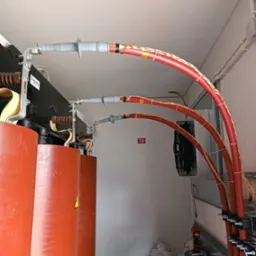 Instalacja elektryczna: trzy czerwone, grube kable podłączone do transformatora, zabezpieczone szarymi osłonami. Widoczne narzędzia i wentylator w tle.