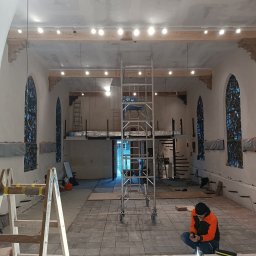 VIKTORISERVIS Stanisław Bohodukh - Wnętrze w trakcie remontu: widoczne rusztowanie, zamontowane oświetlenie i podłoga z płytek. Pracownik w pomarańczowej kurtce sprawdza coś na telefonie.