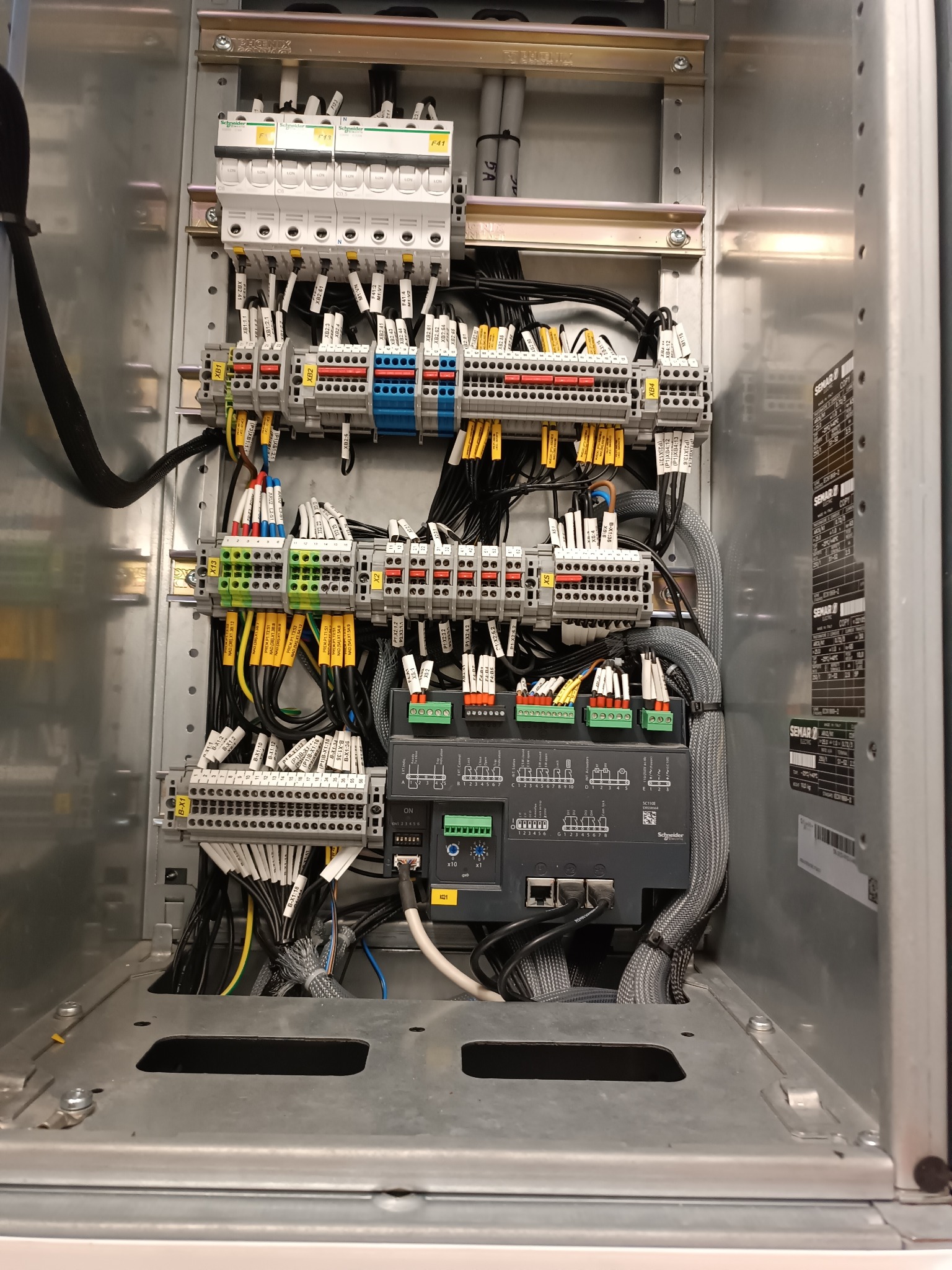 Rozdzielnica elektryczna w metalowej obudowie z licznymi przewodami, złączkami i modułami,  oznaczona etykietami. Widoczne elementy marki Schneider Electric.