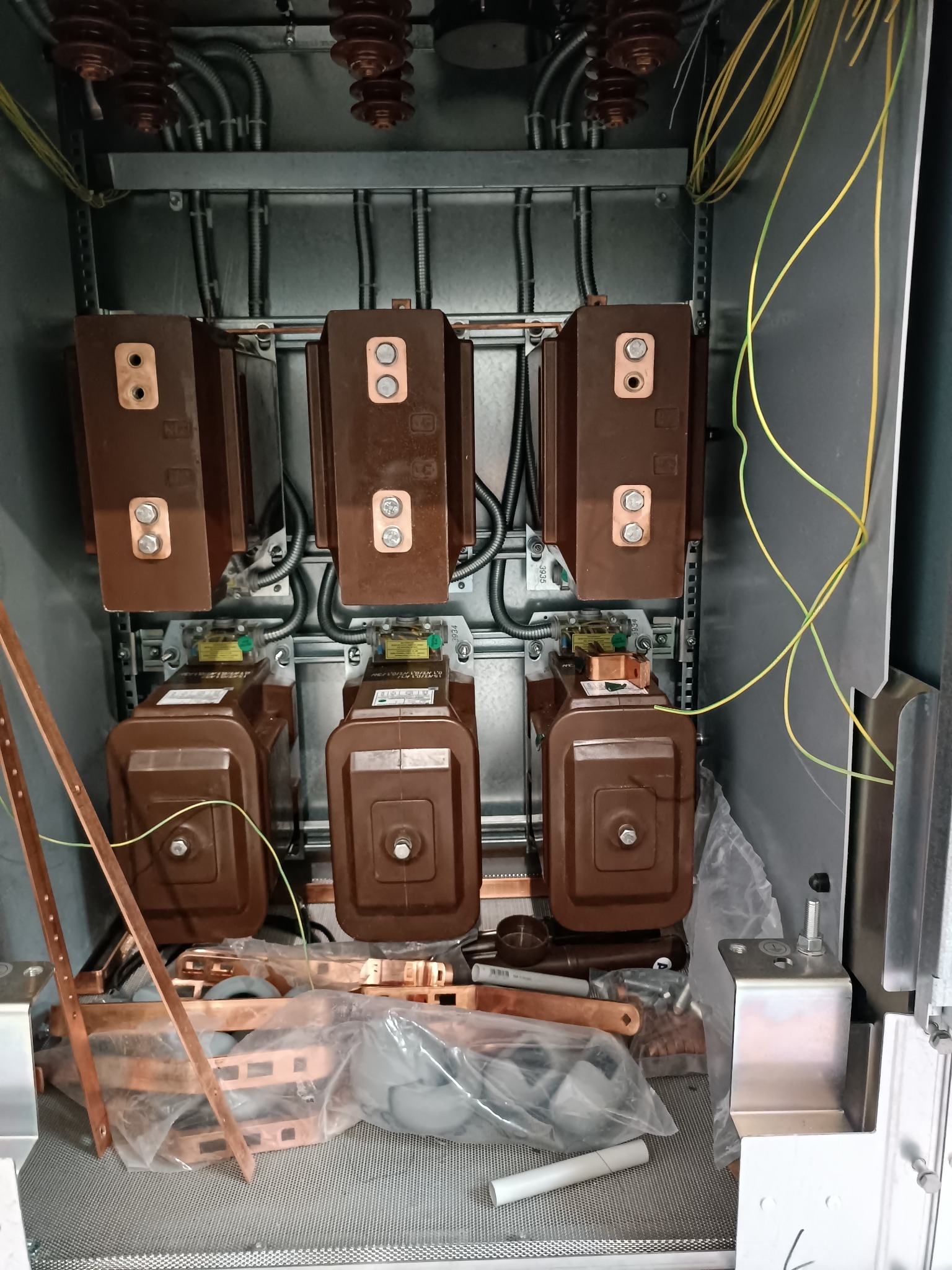 Rozdzielnica elektryczna w trakcie montażu: widoczne brązowe transformatory, kable i miedziane elementy. Częściowo widoczne narzędzia i elementy montażowe.