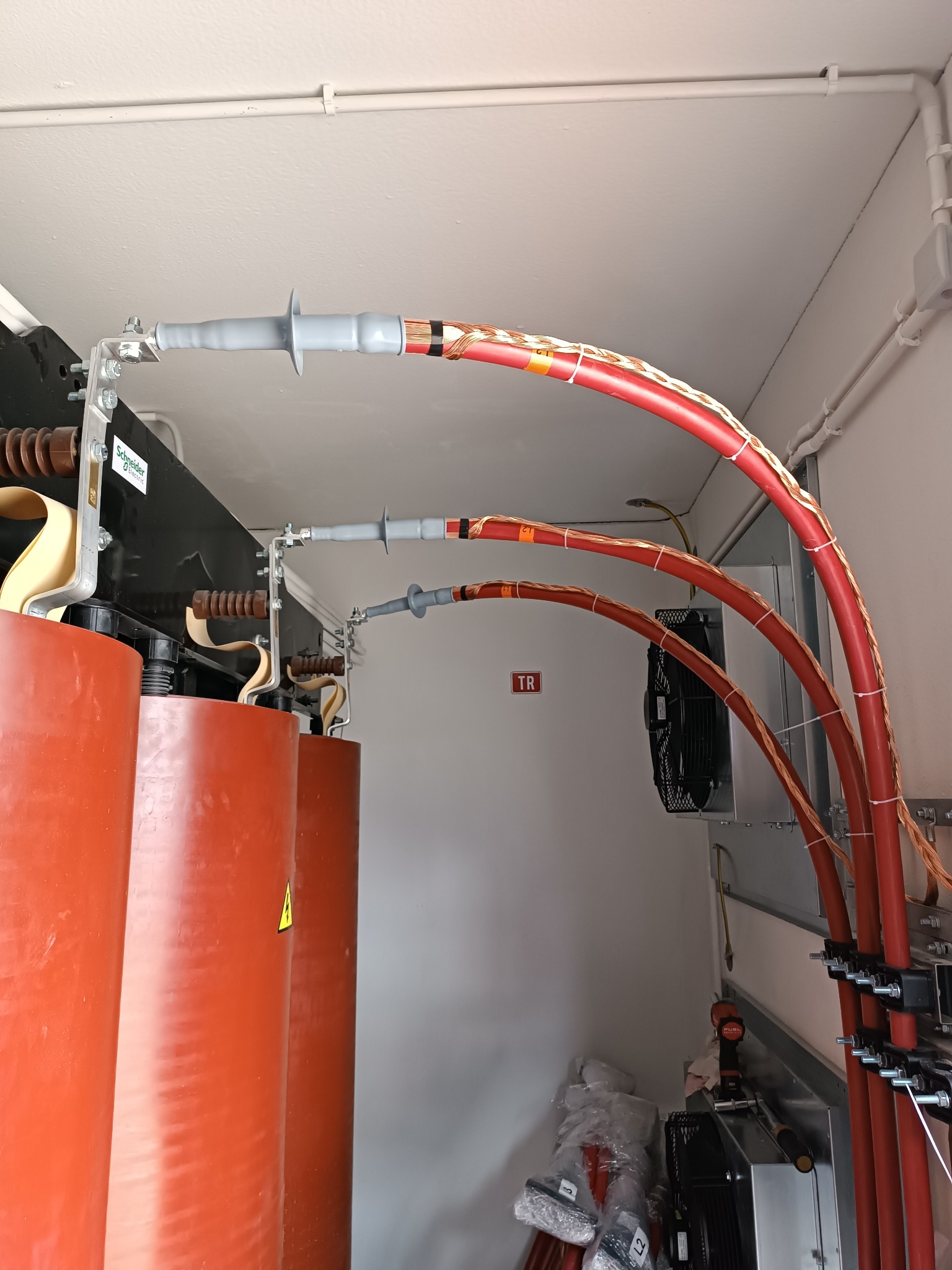 Instalacja elektryczna: trzy czerwone, grube kable podłączone do transformatora, zabezpieczone szarymi osłonami. Widoczne narzędzia i wentylator w tle.