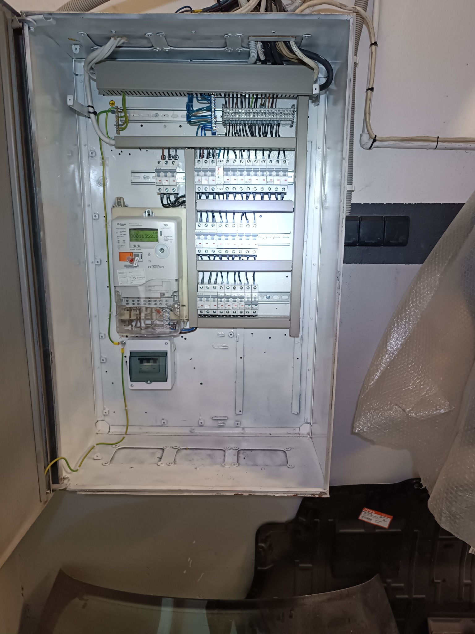 Otwarta, biała skrzynka elektryczna z licznikiem i bezpiecznikami. Widoczne kable i przewody. Elementy instalacji elektrycznej wewnątrz obudowy.